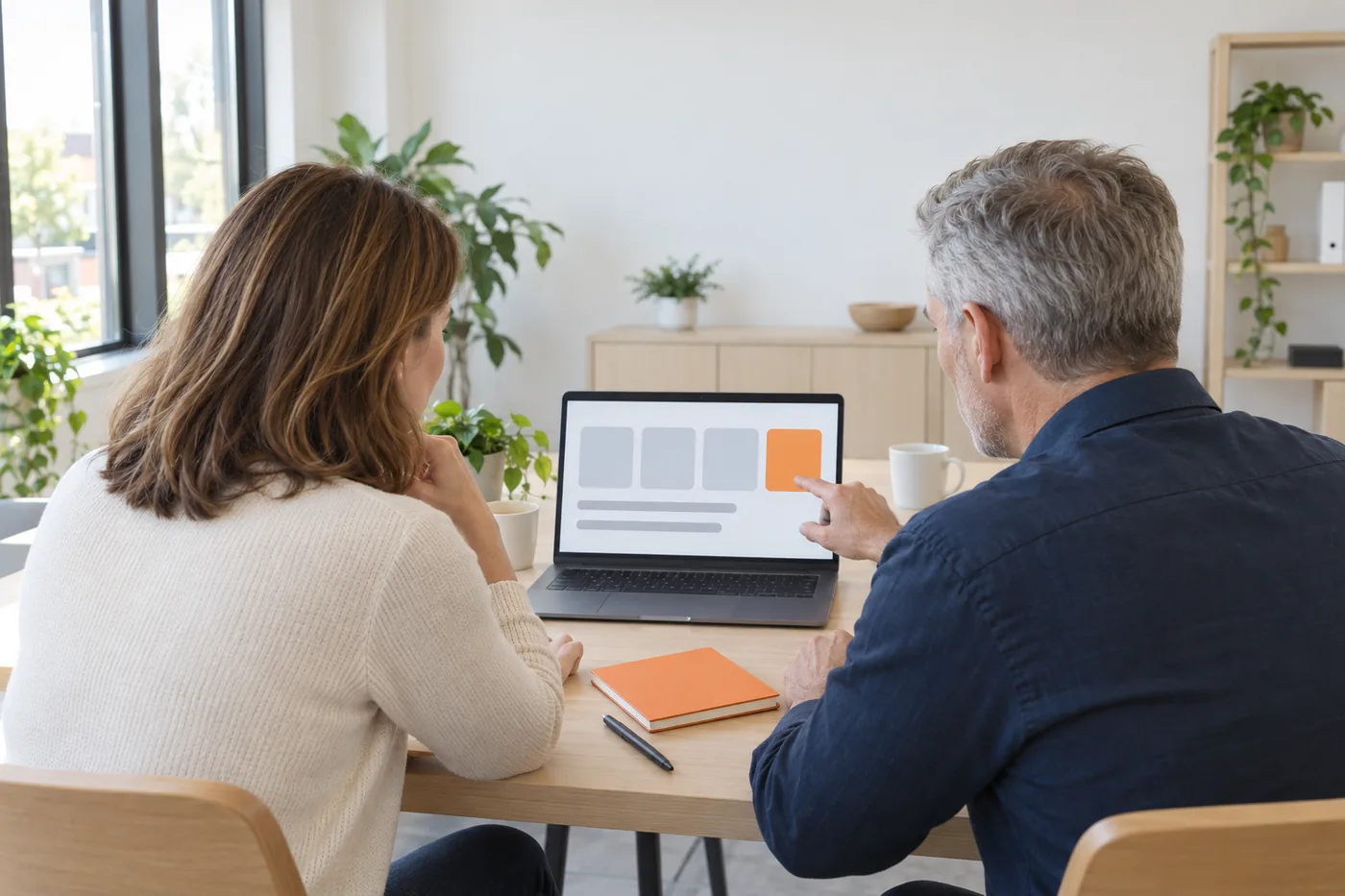 A woman in a cream sweater and a grey-haired man in a navy shirt sit at a wooden table, seen from behind, as he points at an orange card beside three grey cards on a laptop screen.