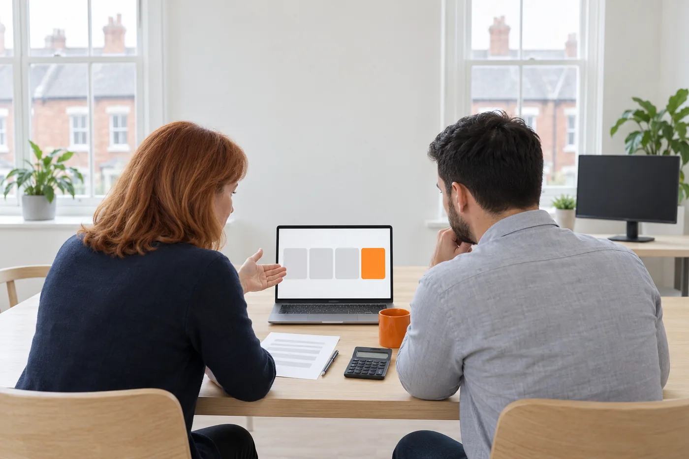 Two people seen from behind at a pale oak table look at a laptop showing three grey cards and one orange card, with a calculator, a printed page and an orange mug beside it and a brick terrace outside the window