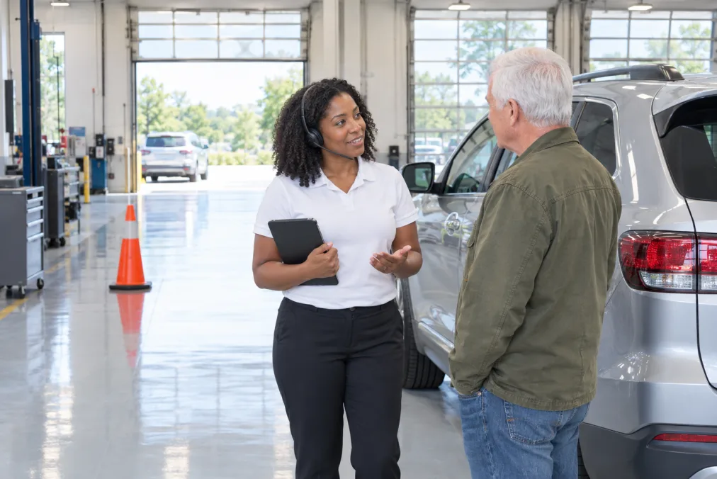 A service advisor wearing a headset and holding a tablet, talking with a customer beside a silver SUV in a dealership service lane