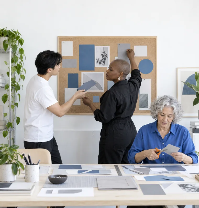 A woman pinning paper cut-outs of page sections onto a cork board while a man hands her the next piece and another woman cuts paper