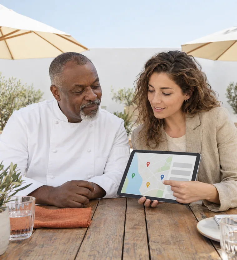 A cook in a white jacket and a woman in a linen blazer looking at a tablet that shows a map with pins on a restaurant terrace
