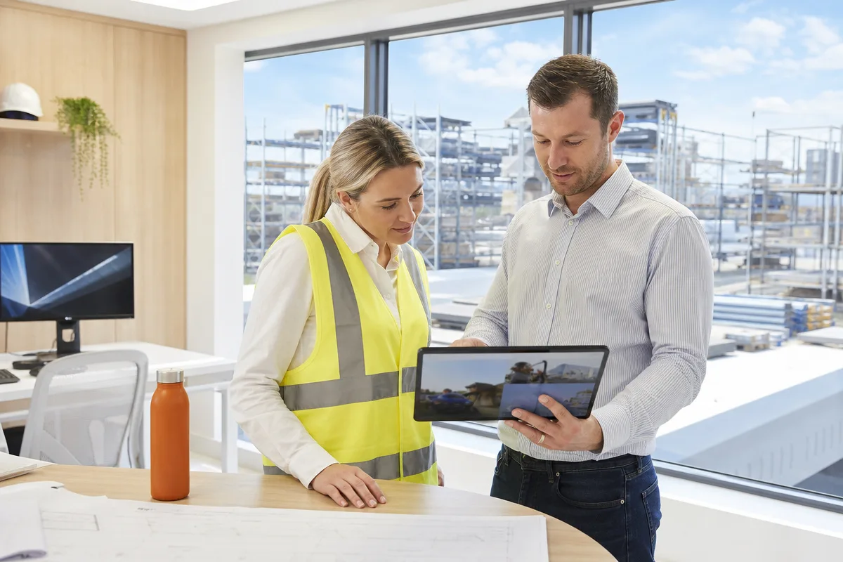 A site manager in a high-visibility vest and a colleague reviewing project photos on a tablet in an industrial site office.