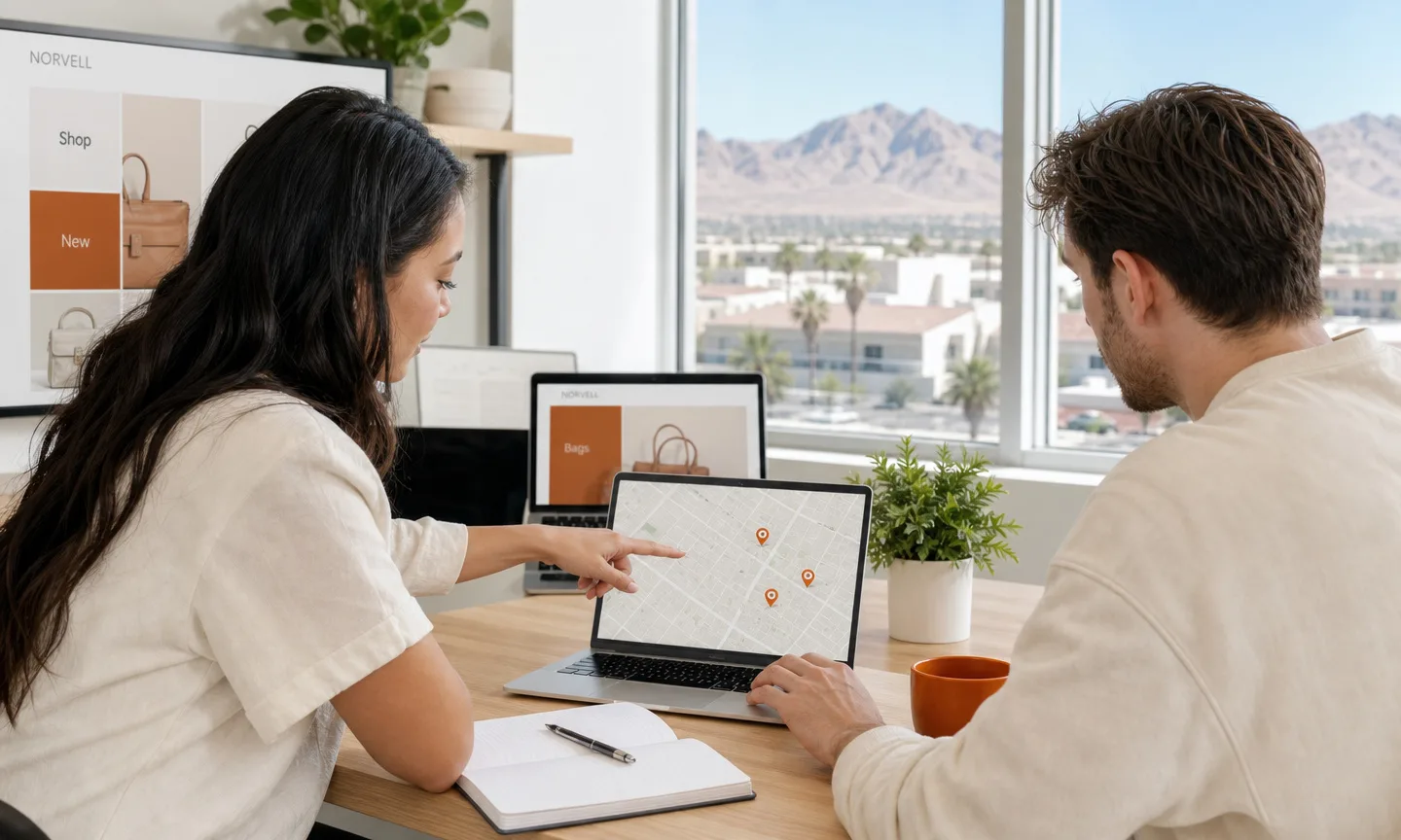 Two marketing specialists reviewing a street map with location pins on a laptop in a bright Las Vegas office with desert mountains through the window